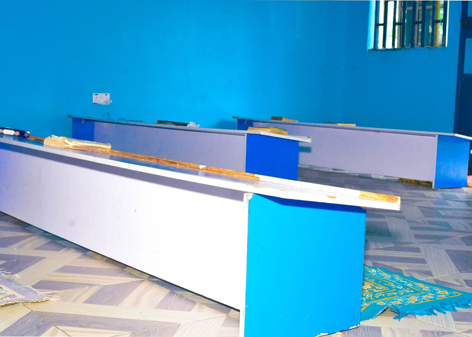 A bright blue hall with rows of desks and prayer mats