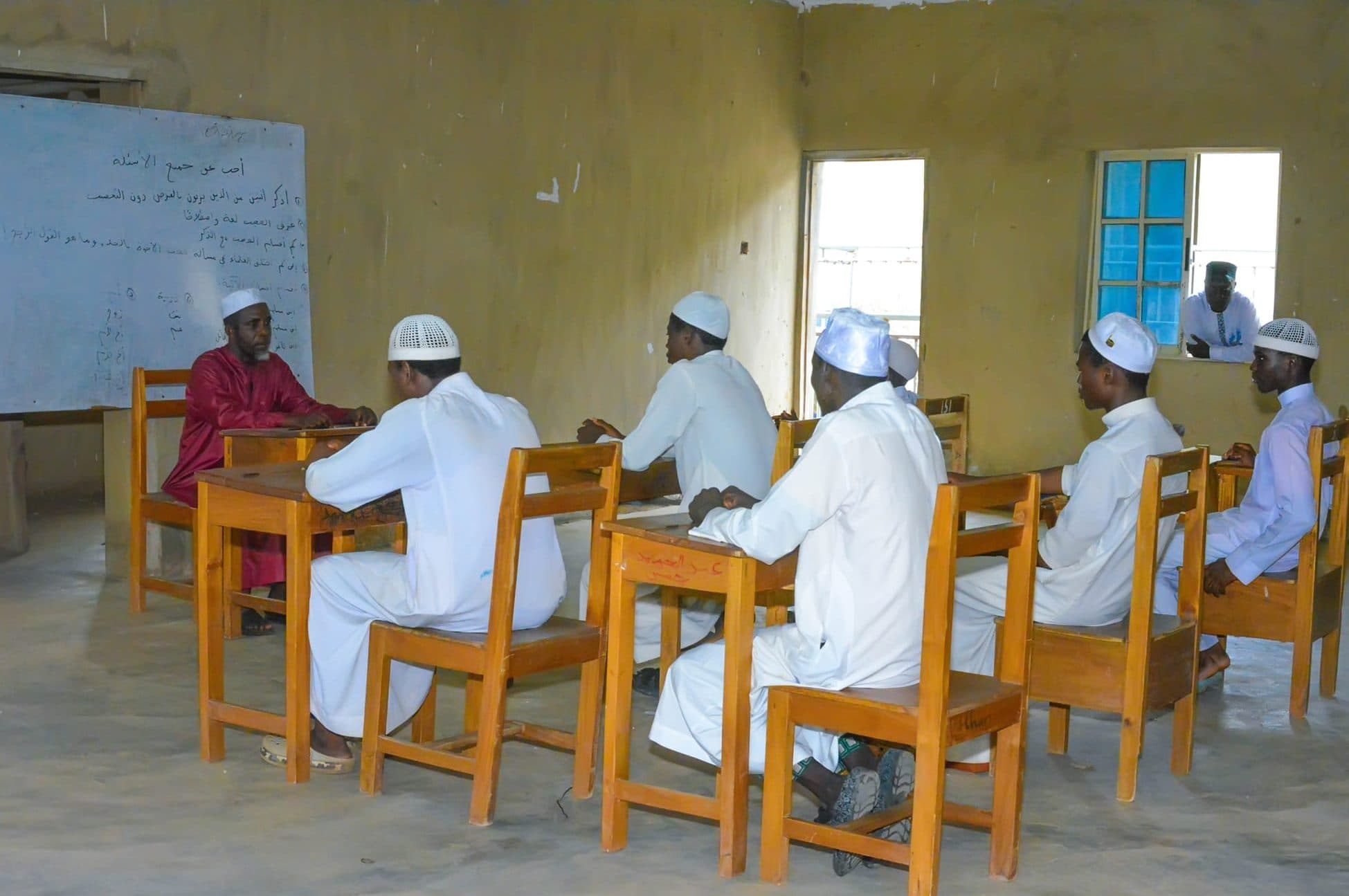 A teacher seated before a whiteboard of Arabic text with older students in white robes and caps